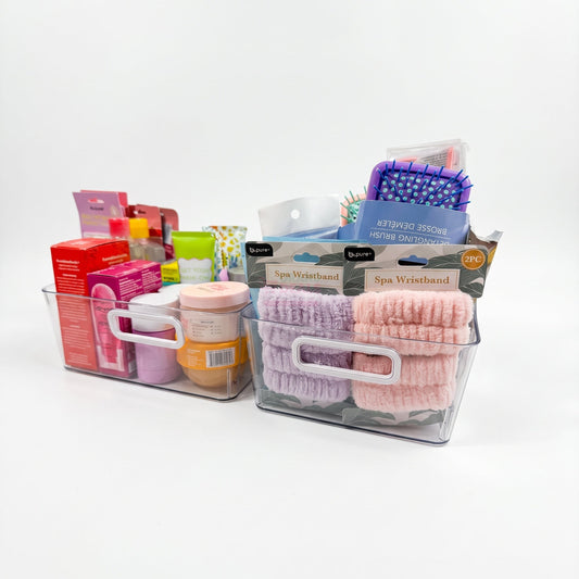 Two clear plastic storage bins with white handles, filled with assorted bath and beauty items including pink and purple spa wristbands, a purple hairbrush, and boxed products, photographed on a clean white background.